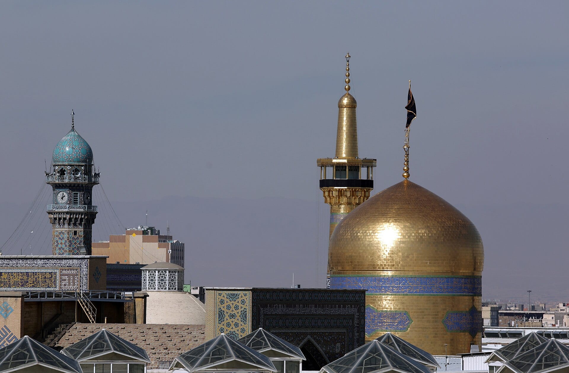 گنبد طلایی و گلدسته زرین حرم امام رضا در کنار مناره فیروزهای در روز