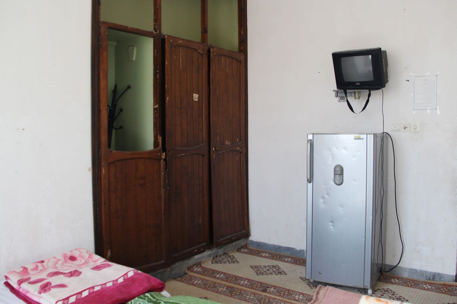 یخچال و تلویزیون کنار درِ چوبی و رختخواب کف اتاق در مسافرخانه آیدین قشم