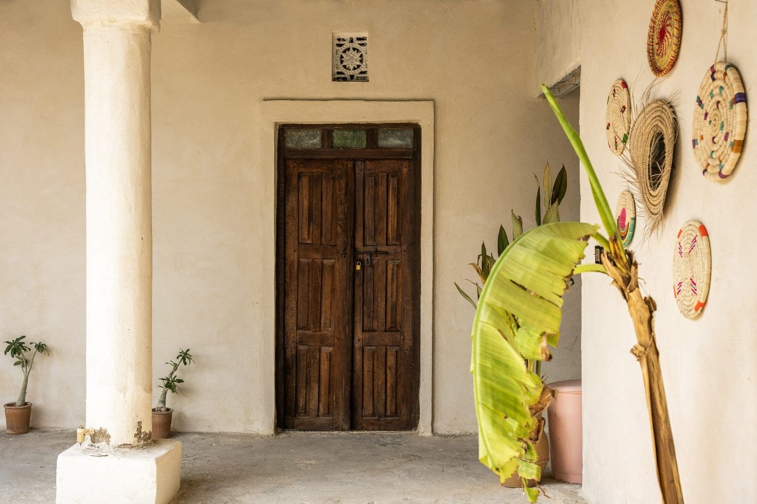 درِ چوبی سنتی و ایوان ستون‌دار با صنایع دستی حصیری روی دیوار در بوم‌گردی خانه مانگرو قشم