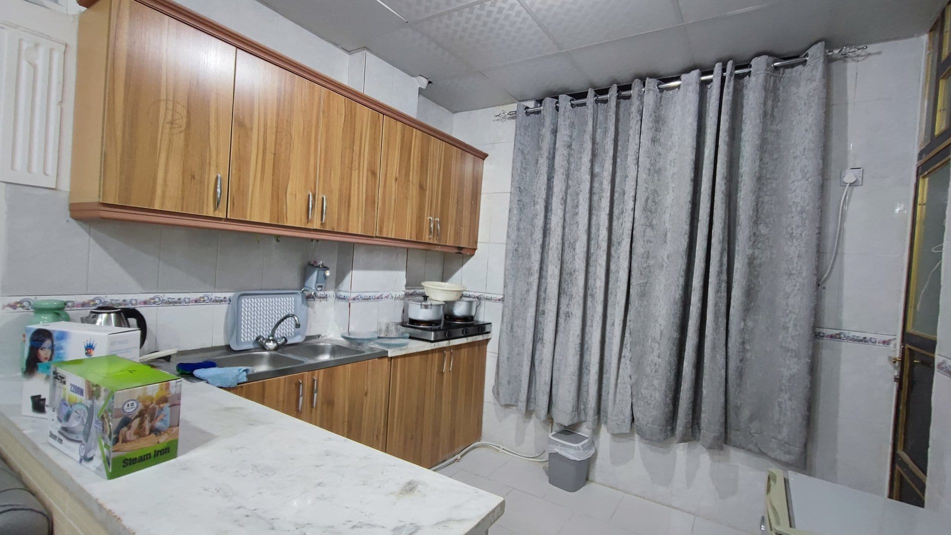 آشپزخانه با کابینت چوبی و پردهٔ طوسی در آپارتمان یک‌خوابه دانشمند ۵ قشم