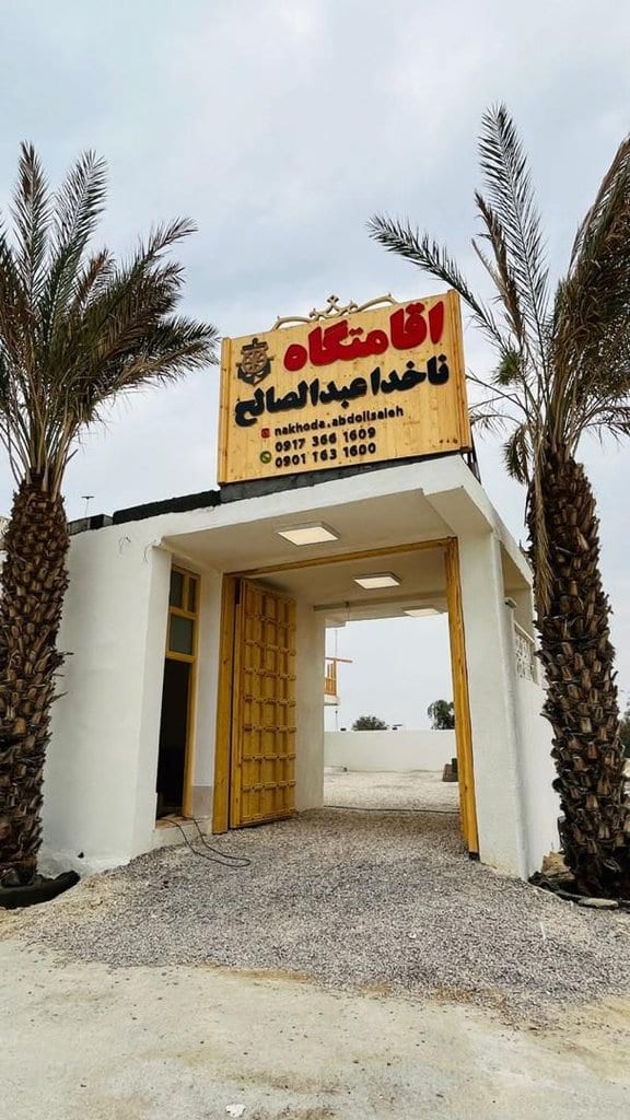 درِ ورودی چوبی باز و سردرِ اقامتگاه بوم‌گردی ناخدا عبدالصالح قشم میان نخل‌ها