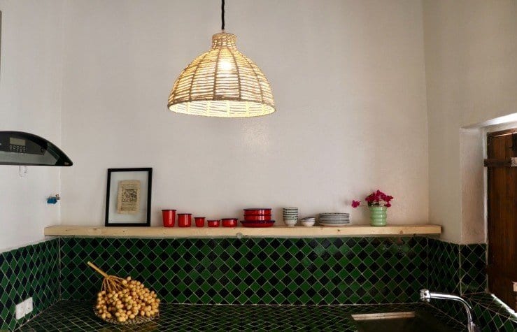 آشپزخانه خانه روشن قشم با کاشی سبز، طبقهٔ چوبی ظروف و چراغ حصیری