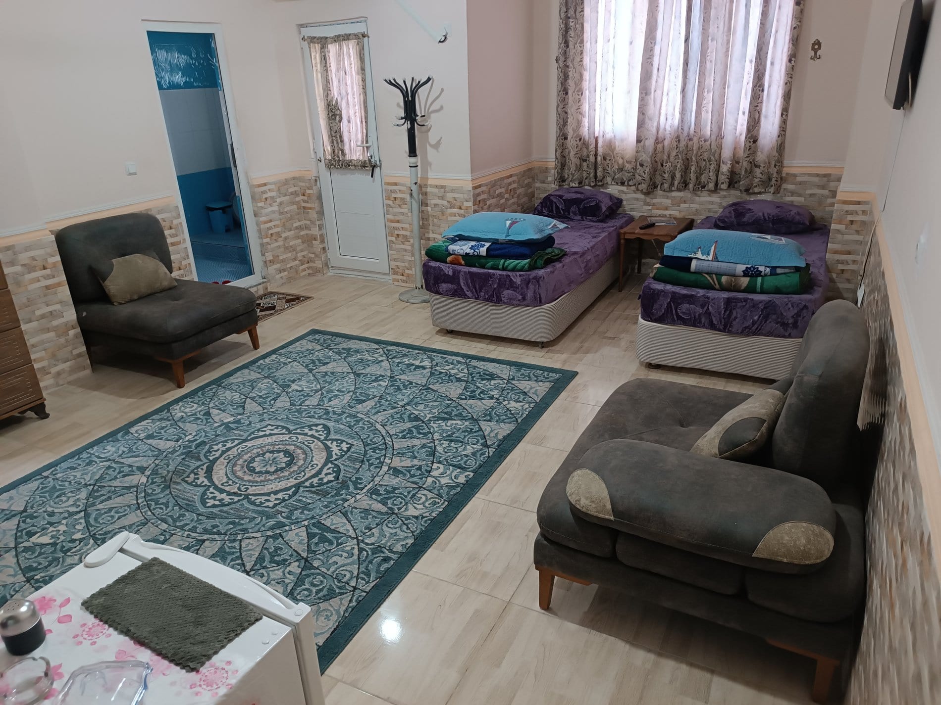 اتاق با دو تخت یک‌نفره و مبل خاکستری کنار فرش مِدالیون‌دار در واحد ۵ برگ سبز قشم