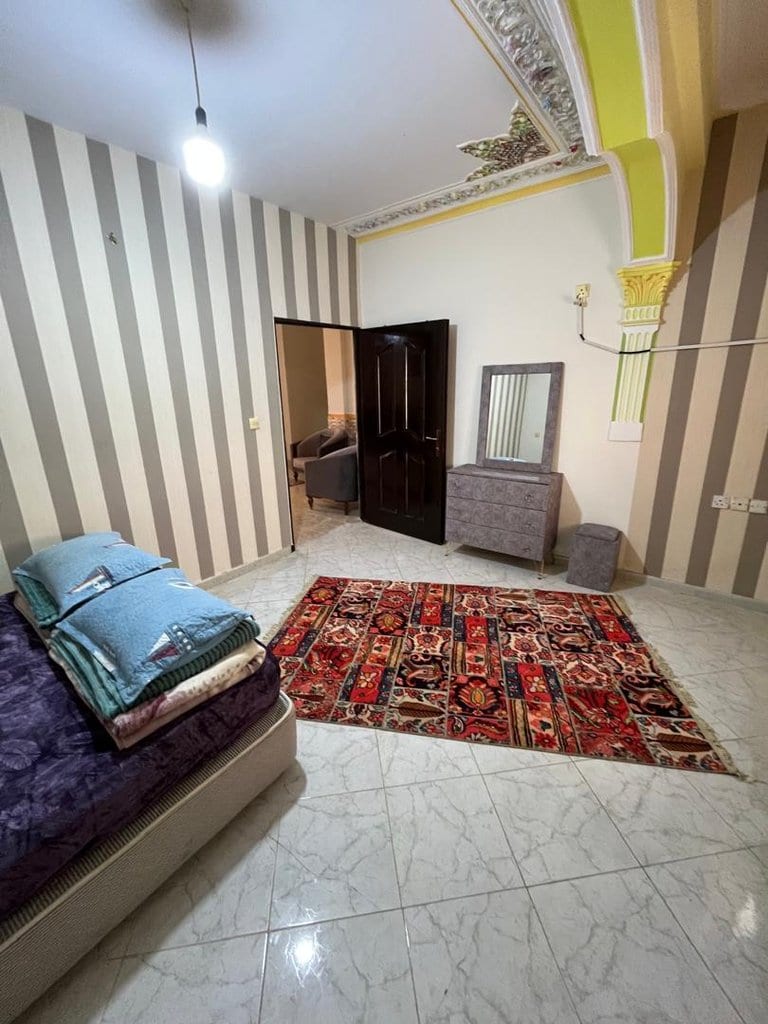 اتاق با دیوار راه‌راه، رخت‌خواب و فرش در واحد دوخوابه برگ سبز قشم